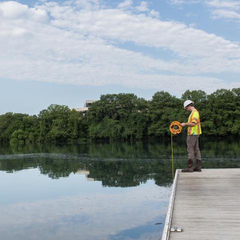 By a body of water, a man, in a hard hat and orange vest, fishes.