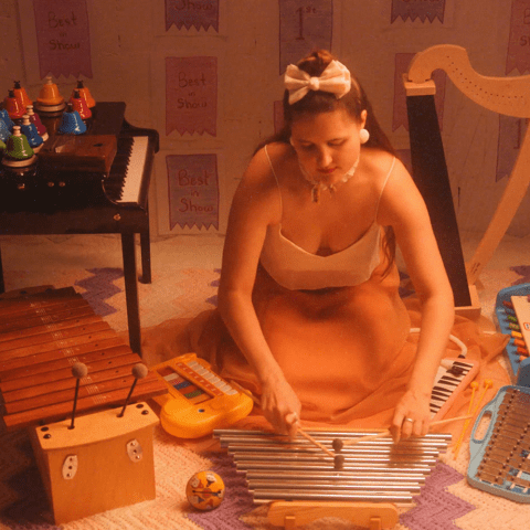 alexa dexa, a queer, non-binary, Sick, Mad, and Disabled white person with light olive skin tones and long golden brown hair, wears a silk gown with a white top and flowing peach skirt and kneels on the floor playing their toy chimes. Surrounding them from left to right are members of their beloved toychestra: a 37-key Schoenhut baby grand toy piano, 20 chromatic desk bells, kalimba, alto xylophone, elephant toy piano, vintage noisemaker, toy glockenspiel, melodica, tap-a-tune xylo-keyboard, and toy harp.