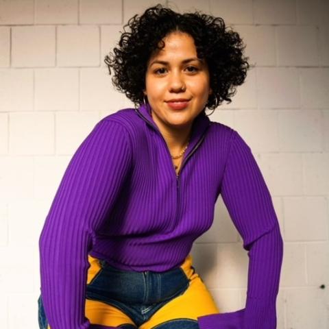 Ali wears a purple sweater and denim pants with yellow cutouts. She has jawlength curly brown hair and she is a Brown woman.