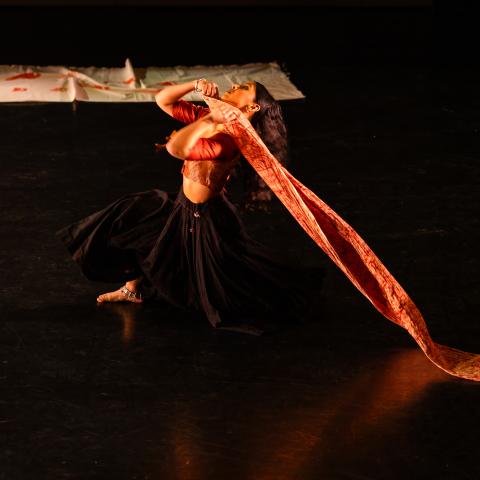 A performer in a dark skirt drags a piece of orange cloth across a stage.