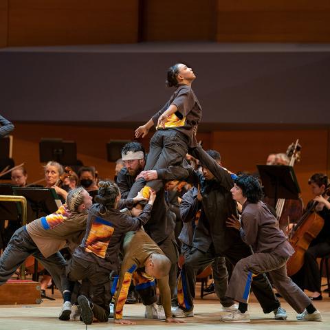 Six dancers hold a woman, who extends her arms, up above them.