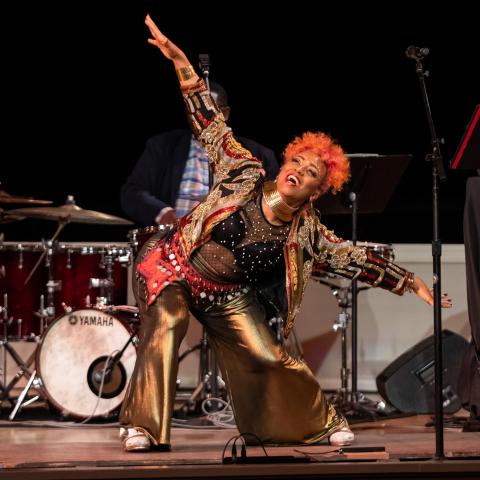 On a stage, between mic stands, a Black woman, with orange hair and gold clothes, bends and holds her arms out wide.