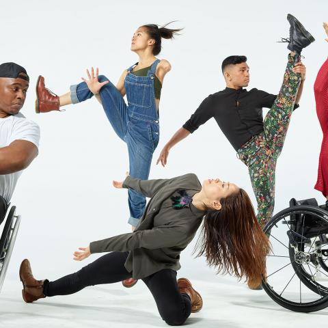 Five dancers, two of which are in wheelchairs, dance in front of a white backdrop.