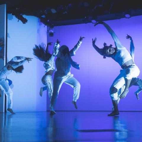 On a stage, with blue lights, seven ladies of color leap and throw their arms around.