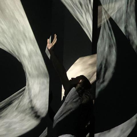 A man dances between screens with projections on them. He holds his hand up.