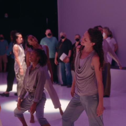In a gallery space, dancers, wearing creams and grays, slouch. A masked crowd watches.