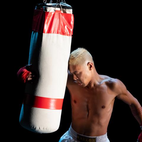 A brown man, with blonde hair, holds a punching bag against his head.