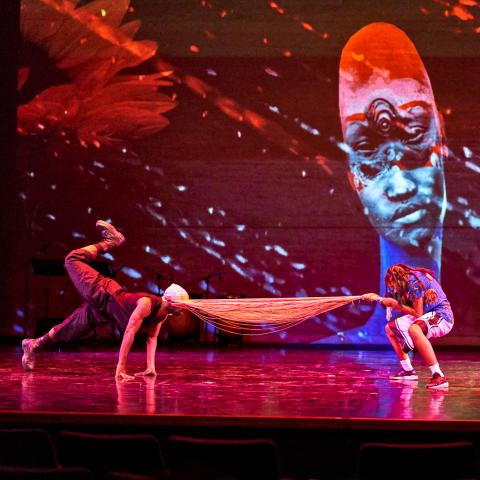 On a stage, a dancer pulls another dancer by their fringe mask, while another looks on. Behind them, a projection of an Afro-Futuristic image, where a person with a long neck and face has three eyes.