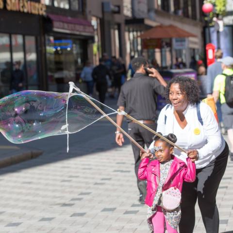 A Black woman and a little Black girl hold a bubble wand with a huge bubble expanding out of the wand.