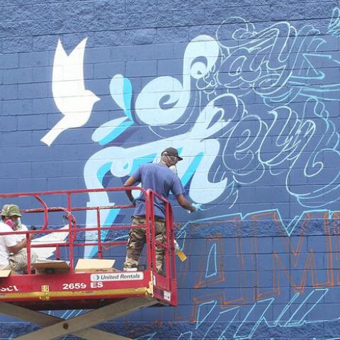 Two folks, on a mechanical lift, paint a mural with text, Say Their Names, and a dove.