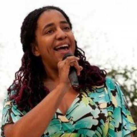 A woman with long hair wearing a bold floral print dress using a cane. She is holding a microphone and sports green Chuck Tayolors.