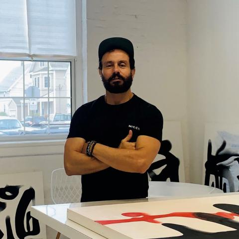 D Derek folds their arms and wears a black baseball cap. They're in an artist studio space with paintings around them.