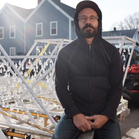 D Derek leans on a teardrop sculpture that is hooked up to a truck for transport. He is backlit by the sun. 