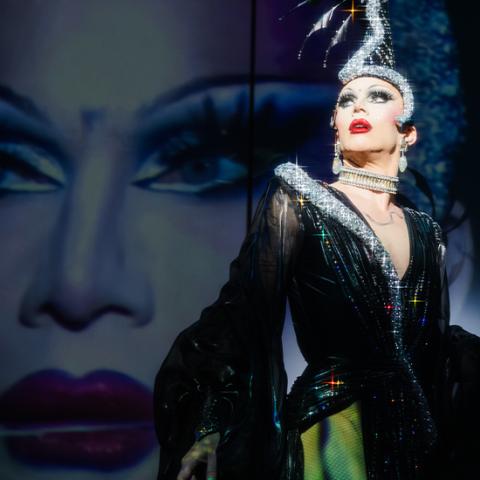 Sasha wears a sheer cloak over pants and a shimmer headpiece that snakes along her scalp. Behind her a projection of her face, which is spotlit.