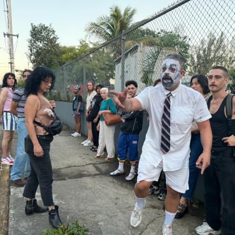 A person whose face has been painted white stands on a street, pointing at something offscreen. Eleven people look in the direction he's pointing.