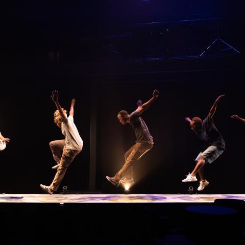 Five performers on a stage are mid leap with their arms raised over their heads and extended outwards.