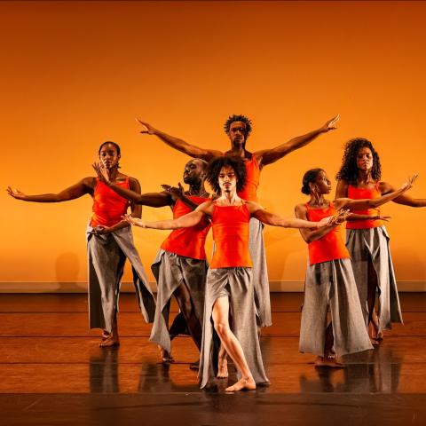 On a stage, with an orange backdrop, six dancers, in orange tank tops and grey skirts, spread their arms.