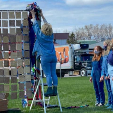 A woman hangs some fabric over a pole that is part of an installation with dangling silver squares. Folks look on as she does this.