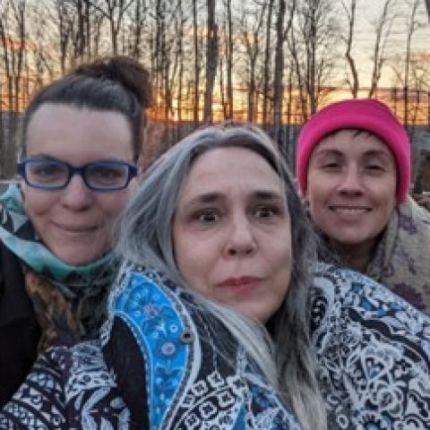 Three white women dressed for chilly weather smile and scrunch together for a selfie as the sun sets through the bare trees behind them 
