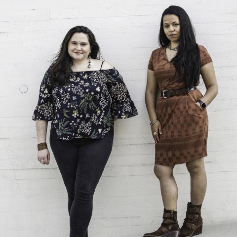 Two women of color pose next to each other on some outdoor steps, in front of white bricks. They both wear heels - one wears a brown wrap dress and brown, heeled boots, while the other wears a flowy black and white top off one shoulder and black denim with grey gladiator sandals.