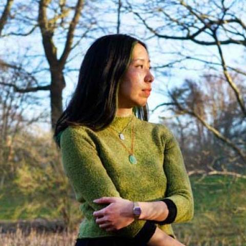 Lily stands in a field of dried branches, holding one arm with another and looking off into the distance. She wears a grass green sweater and a jade necklace on a red string.