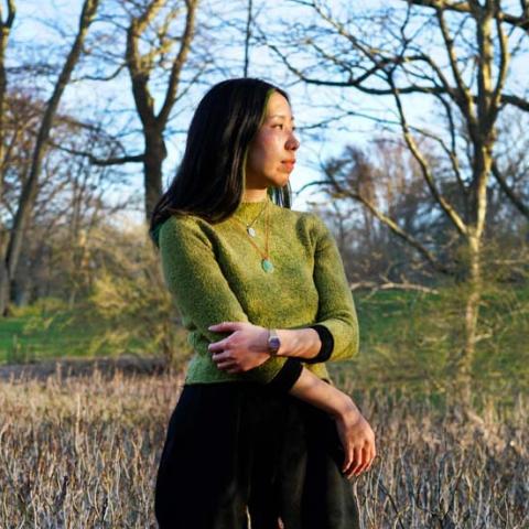 Lily stands in a field of dried branches, holding one arm with another and looking off into the distance. She wears a grass green sweater and a jade necklace on a red string.