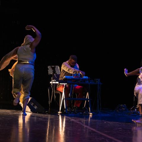 On a stage, Black women dance while a Black man plays the keyboard. Spotlights are also on the stage.