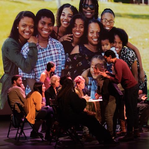 On a stage, six white folks sit around a table. A white man leans over them and gestures with his hands. Behind them is a portrait of a group of people of color.