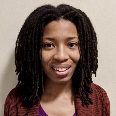 Micah is a black woman with shoulder length braids. She wears a maroon cardigan, indoors.