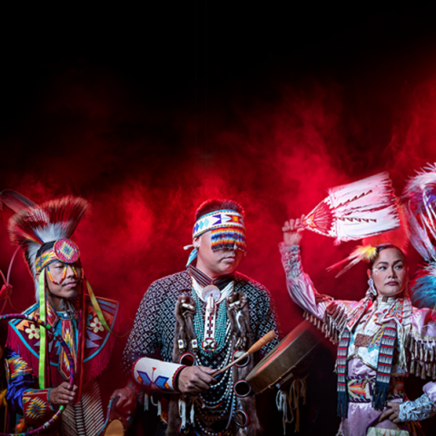In front of red smoke, five performers in traditional native garb dance and play music.