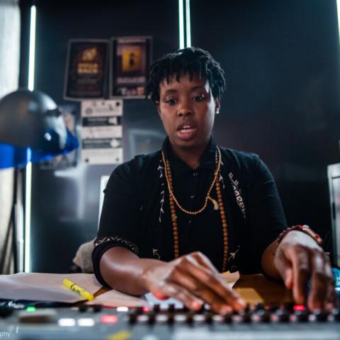 In a small dark space, a Black woman works a sound board.