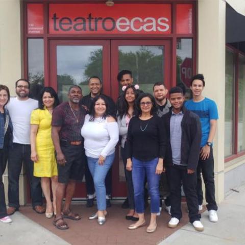 Twelve members of Teatro ECAS stand together on the sidewalk outside of the theater